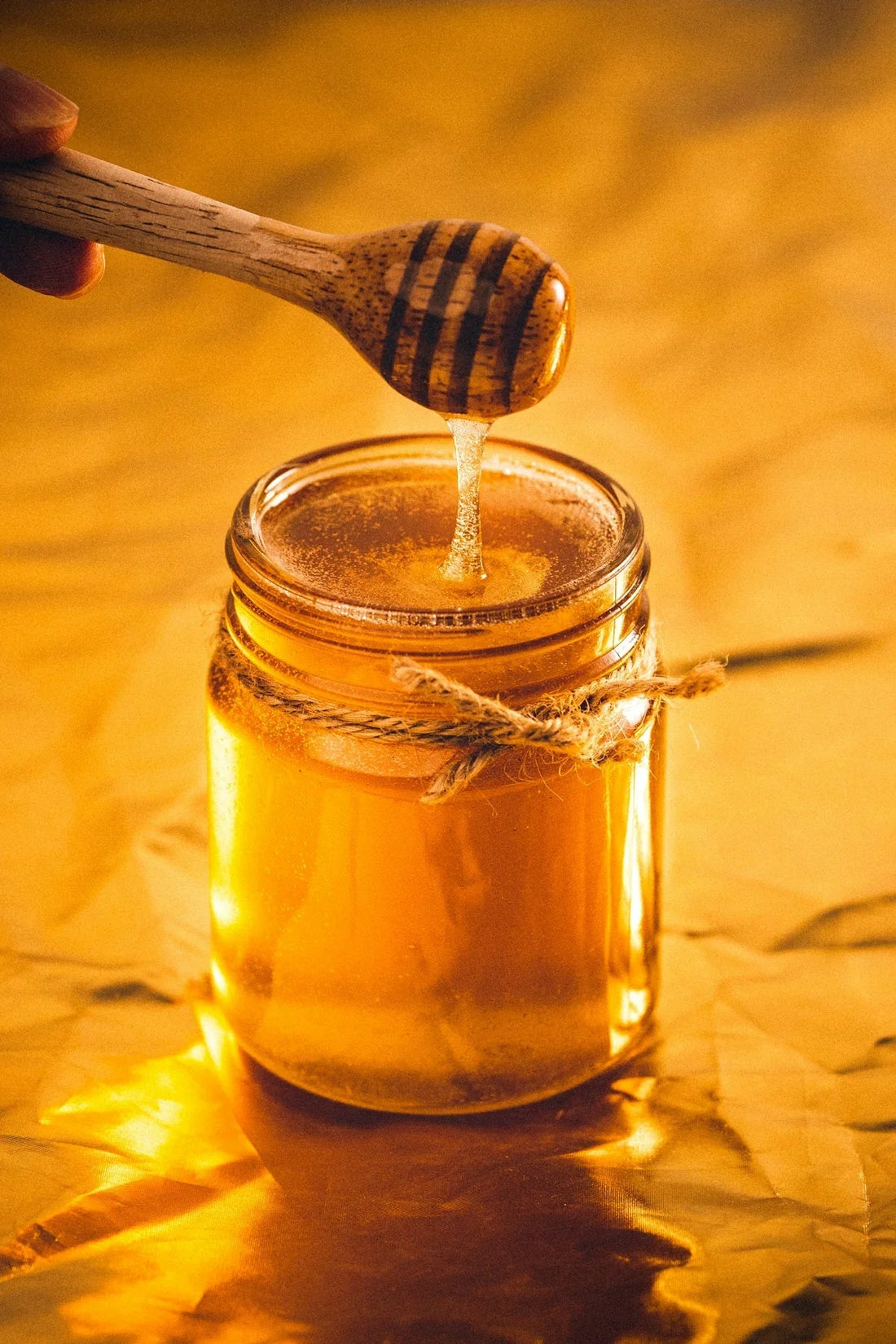 Honey jar with a wooden honey dipper dripping golden honey, tied with twine on a warm background