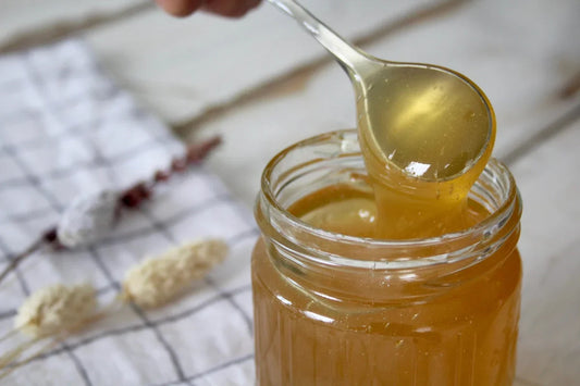 Close-up of honey dripping from a spoon into a glass jar with dried flowers on a checkered cloth