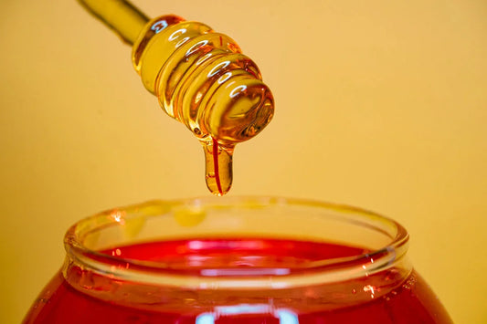 Close-up of honey dipper dripping golden honey into an open glass jar on yellow background